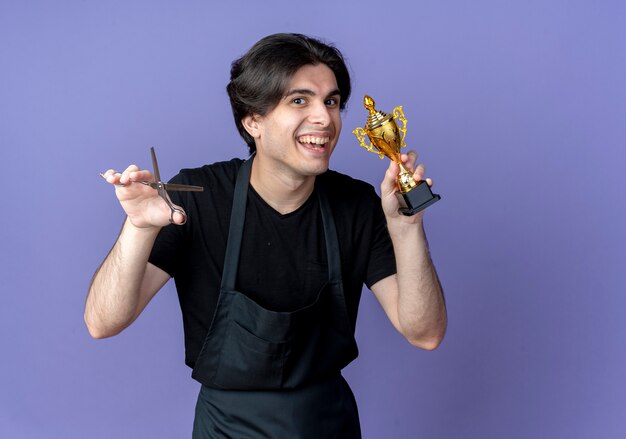 Peluquero masculino guapo joven sonriente en uniforme sosteniendo tijeras y copa ganador aislado en azul