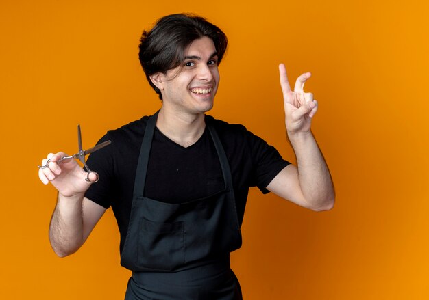 Peluquero masculino guapo joven sonriente en uniforme sosteniendo tijeras y mostrando gesto de paz aislado en naranja