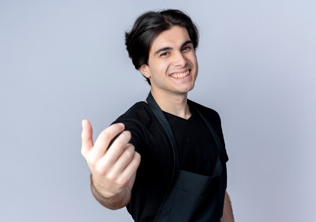 Peluquero masculino guapo joven sonriente en uniforme tendiendo la mano a la cámara aislada en blanco