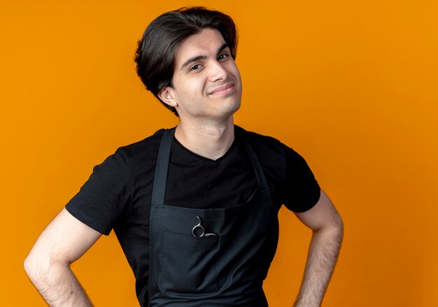 Peluquero masculino hermoso joven complacido en uniforme que pone la mano en la cadera aislada en naranja