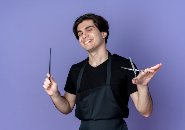 Peluquero masculino hermoso joven sonriente en uniforme que sostiene el peine con las tijeras aisladas en azul