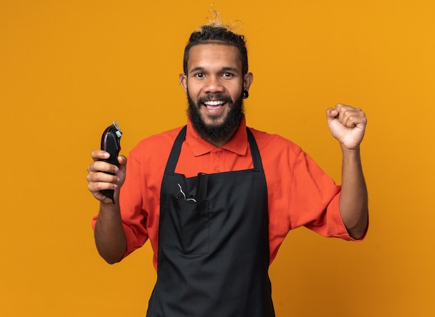 Peluquero masculino joven alegre vistiendo uniforme sosteniendo cortapelos mirando al frente haciendo sí gesto aislado en la pared naranja