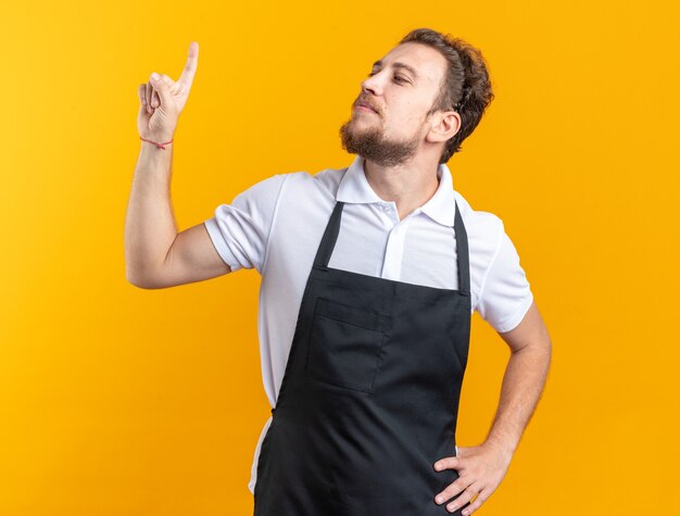 Peluquero masculino joven complacido vistiendo puntos uniformes en poner la mano en la cadera aislada en la pared amarilla