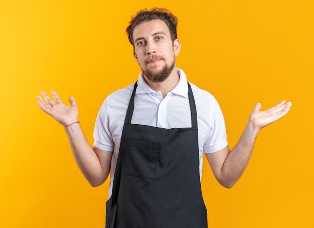 Peluquero masculino joven complacido vistiendo uniforme extendiendo las manos aisladas sobre fondo amarillo