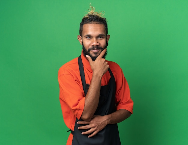 Peluquero masculino joven complacido vistiendo uniforme manteniendo la mano en la barbilla mirando al frente aislado en la pared verde con espacio de copia