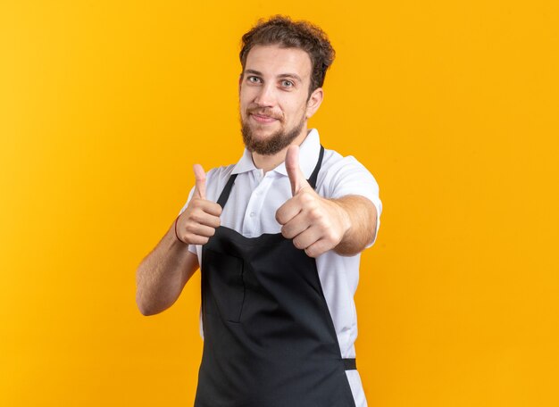 Peluquero masculino joven complacido vistiendo uniforme mostrando Thumbs up aislado sobre fondo amarillo