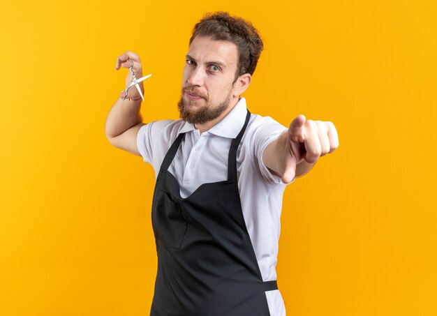 Peluquero masculino joven complacido vistiendo uniforme sosteniendo puntos de tijeras en la cámara aislada en la pared amarilla