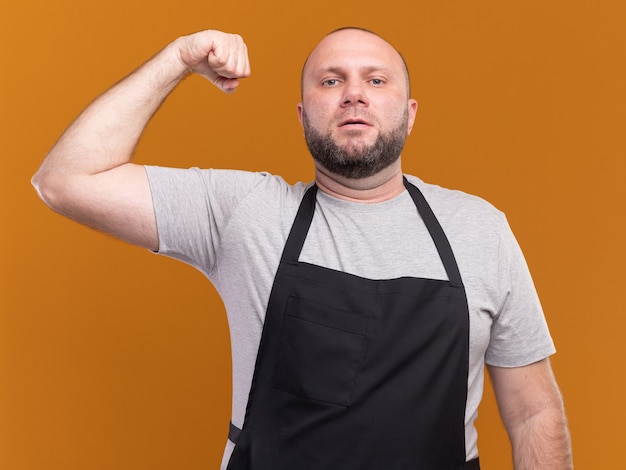 Peluquero masculino de mediana edad eslavo confiado en uniforme haciendo gesto fuerte aislado en la pared naranja