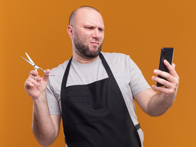 Peluquero masculino de mediana edad eslavo disgustado en uniforme sosteniendo tijeras y mirando el teléfono en la mano aislado en la pared naranja