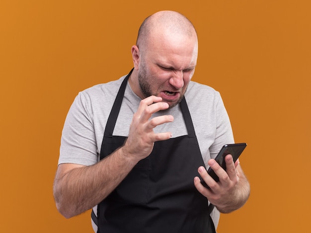 Peluquero masculino de mediana edad eslavo enojado en uniforme sosteniendo y mirando el teléfono aislado en la pared naranja