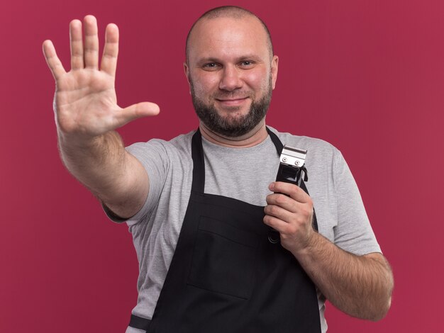 Peluquero masculino de mediana edad eslavo sonriente en uniforme sosteniendo cortapelos mostrando cinco aislados en la pared rosa