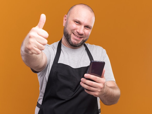 Peluquero masculino de mediana edad eslavo sonriente en uniforme sosteniendo el teléfono mostrando el pulgar hacia arriba aislado en la pared naranja