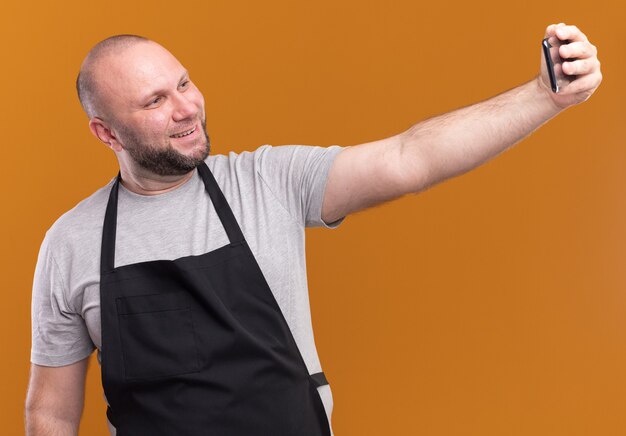 Peluquero masculino de mediana edad eslavo sonriente en uniforme toma un selfie aislado en la pared naranja