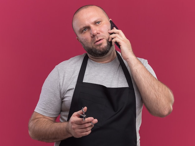 Peluquero masculino de mediana edad eslavo en uniforme sosteniendo tijeras - aislado en la pared rosa