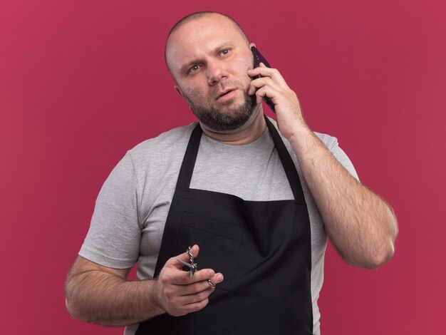 Peluquero masculino de mediana edad eslavo en uniforme sosteniendo tijeras - aislado en la pared rosa