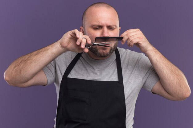 Peluquero masculino de mediana edad seguro en el bigote de corte uniforme con tijeras aisladas en la pared púrpura