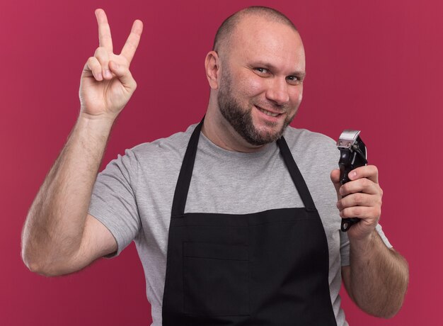 Peluquero masculino de mediana edad sonriente en uniforme sosteniendo cortapelos mostrando gesto de paz aislado en la pared rosa