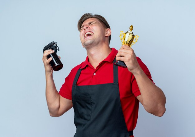 Peluquero masculino rubio alegre joven en uniforme sostiene las herramientas del peluquero y la taza del ganador mirando hacia arriba aislado en el espacio en blanco con espacio de copia