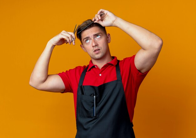 Peluquero masculino rubio ansioso joven en uniforme tratando de cortar su cabello con peine y tijeras aislado en espacio naranja con espacio de copia