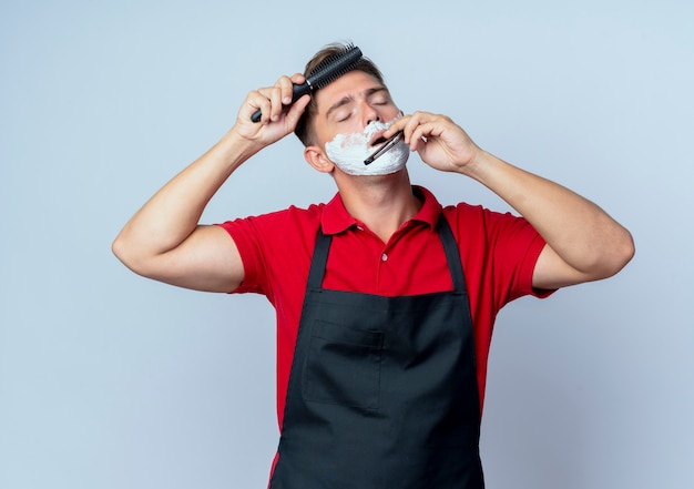 Peluquero masculino rubio complacido joven en la cara manchada uniforme con la espuma de afeitar que sostiene el pelo recto que peina de la maquinilla de afeitar aislado en el espacio en blanco con espacio de la copia