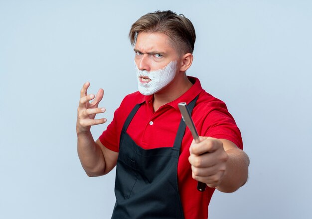 Peluquero masculino rubio molesto joven en cara manchada uniforme con espuma de afeitar sosteniendo navaja de afeitar aislada en el espacio en blanco con espacio de copia