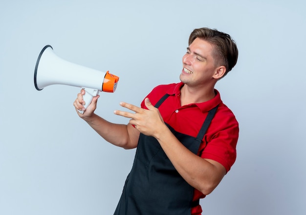 Peluquero masculino rubio sonriente joven en uniforme sostiene altavoz mirando al lado aislado sobre fondo blanco con espacio de copia