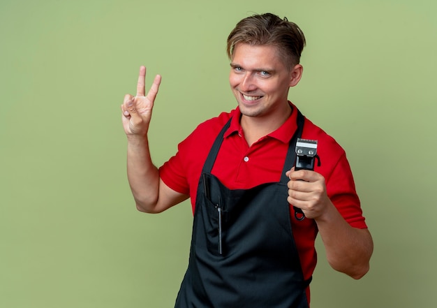 Peluquero masculino rubio sonriente joven en uniforme sostiene el cortapelos y hace gestos signo de mano de victoria aislado en el espacio verde oliva con espacio de copia