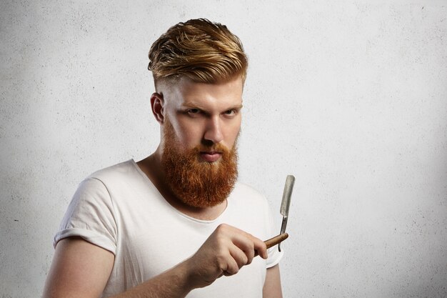 Peluquero pelirrojo de moda con corte de pelo elegante y barba difusa sosteniendo una navaja de afeitar en sus manos, con expresión seria.