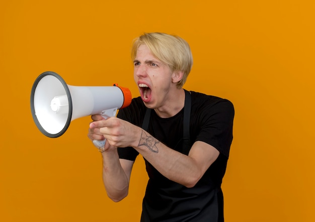 Foto gratuita peluquero profesional en delantal gritando al megáfono en voz alta con expresión agresiva de pie sobre la pared naranja