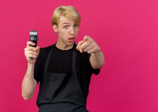 Peluquero profesional en delantal sosteniendo trimmer pointign con el dedo índice a la cámara con expresión escéptica