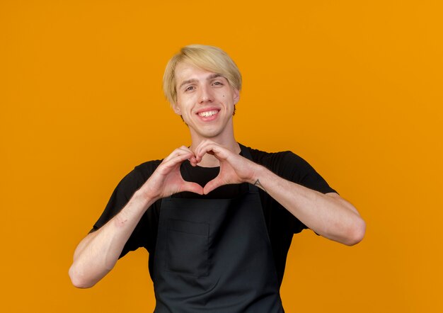 Peluquero profesional hombre en delantal haciendo gesto de corazón con los dedos mirando al frente sonriendo alegremente feliz y positivo de pie sobre la pared naranja