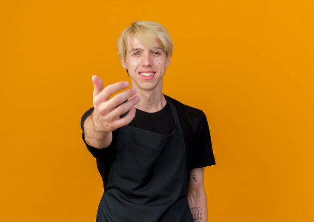 Peluquero profesional hombre en delantal haciendo gesto de ven aquí con la mano sonriente de pie sobre la pared naranja