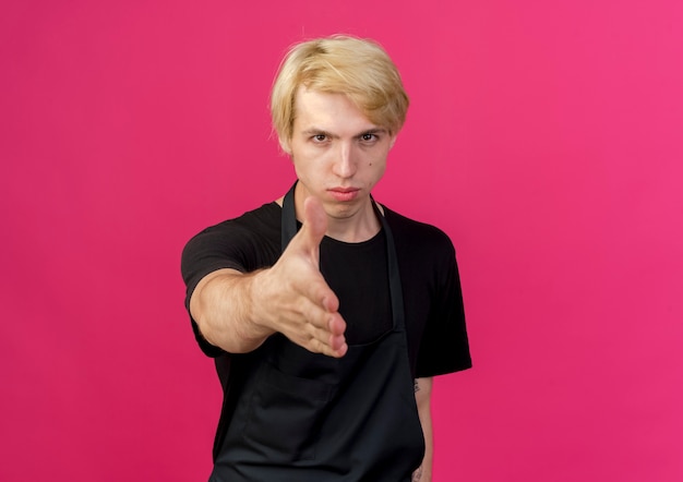Peluquero profesional hombre en delantal mirando al frente con rostro serio ofreciendo gesto de saludo de mano de pie sobre la pared rosa