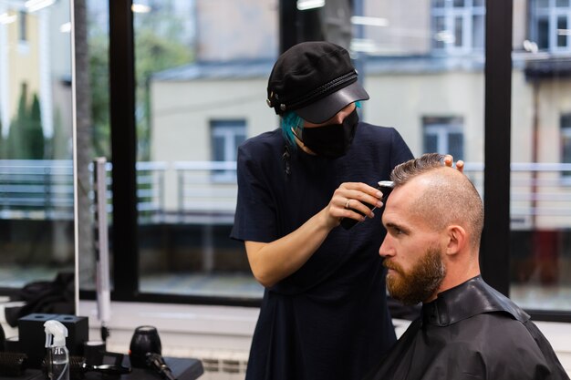 Peluquero profesional con mascarilla protectora, corte de pelo para hombre brutal barbudo europeo en salón de belleza