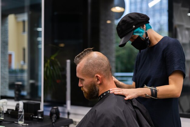 Peluquero profesional con mascarilla protectora, corte de pelo para hombre brutal barbudo europeo en salón de belleza