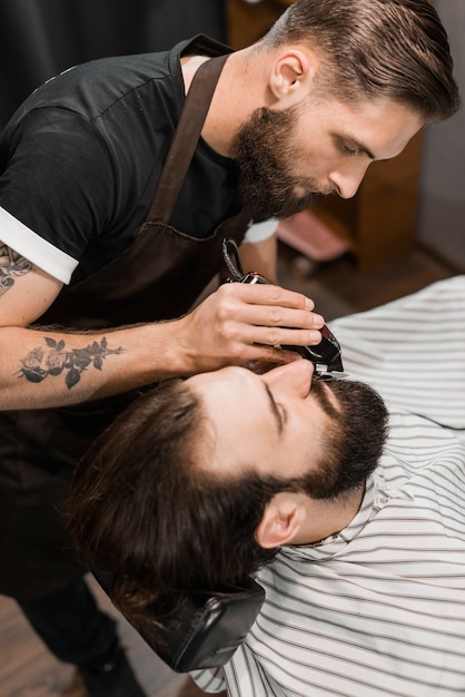 Peluquero profesional que estiliza la barba del hombre con el condensador de ajuste eléctrico