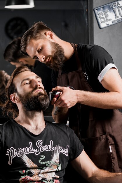 Peluquero que corta la barba del hombre con la maquinilla de afeitar eléctrica