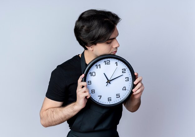 Peluquero de sexo masculino hermoso joven en uniforme que sostiene el reloj de pared alrededor de la cara aislada en blanco