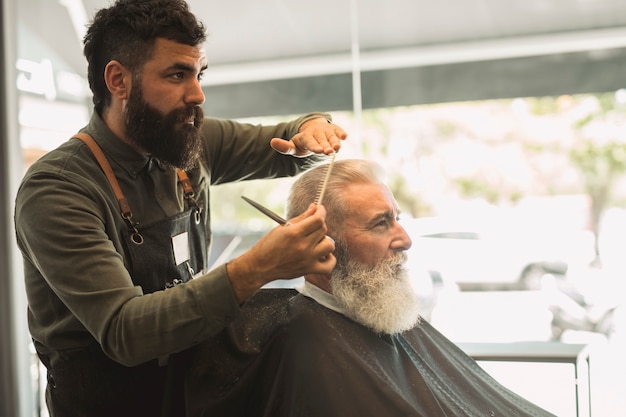 Peluquero de sexo masculino que peina el pelo del cliente mayor en barbería