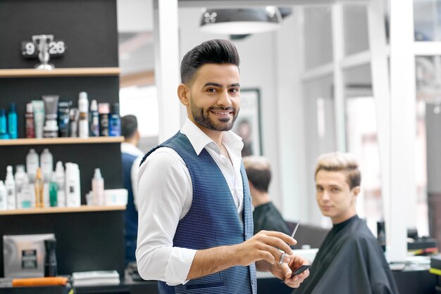 Peluquero sonriente parado cerca de un cliente joven sentado cerca del espejo