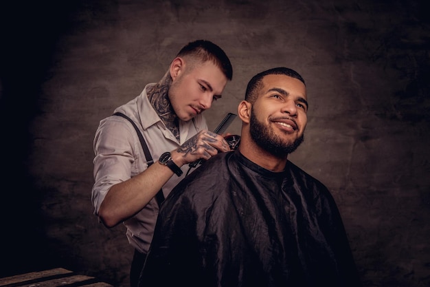 El peluquero tatuado profesional a la antigua le hace un corte de pelo a un cliente afroamericano, usando una recortadora y un peine. Aislado sobre fondo oscuro con textura.