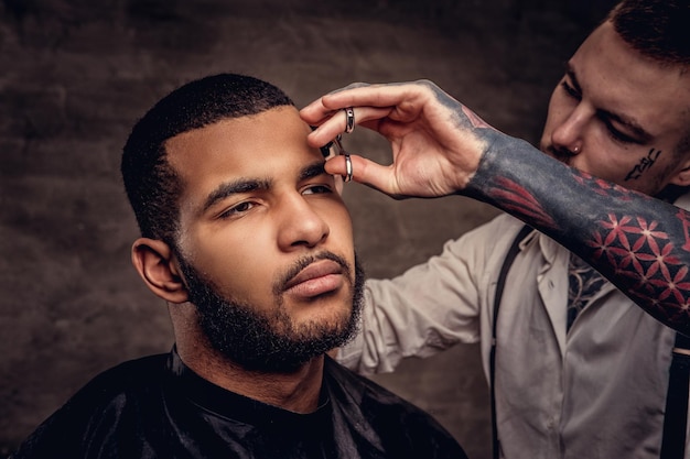 El peluquero tatuado profesional a la antigua le hace un corte de pelo a un cliente afroamericano, usando tijeras y peine. Aislado sobre fondo oscuro con textura.