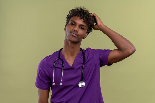 Un pensamiento joven apuesto médico de piel oscura con cabello rizado vistiendo uniforme violeta con estetoscopio sosteniendo la mano en la cabeza mientras está en un espacio verde
