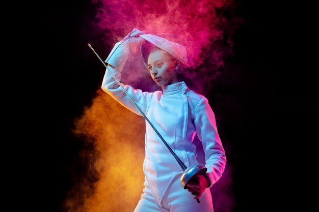 Pensamientos del ganador. Chica adolescente en traje de esgrima con espada en mano humo de neón aislado iluminado. Practicar el entrenamiento en movimiento, acción. Copyspace. Deporte, juventud, estilo de vida saludable.