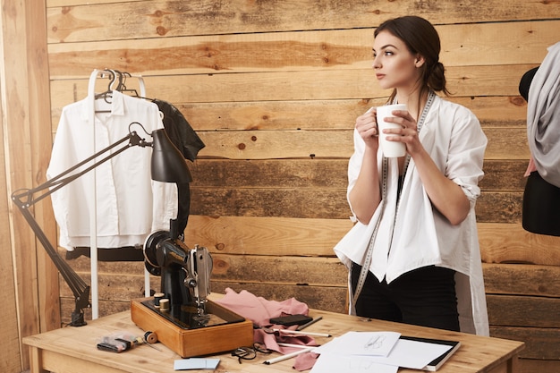 Los pensamientos me llevan lejos. Joven diseñador de ropa linda de pie en el taller, teniendo un descanso de la costura, tomando café y pensando mientras mira a un lado, planeando un nuevo diseño de prenda