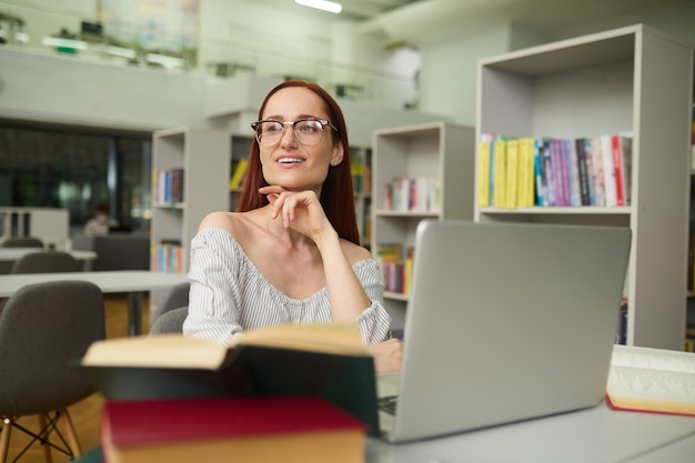 Pensamientos positivos. Mujer joven sonriente de pelo largo con gafas sentada en la mesa con una computadora portátil y libros mirando hacia un lado tocando la mano con la barbilla en una habitación iluminada