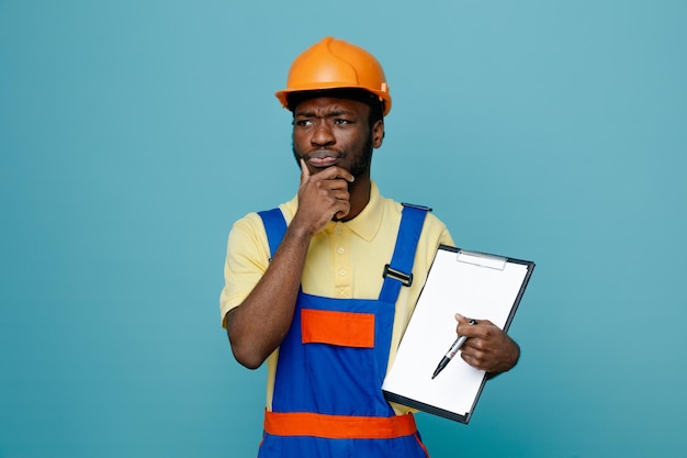 Pensando agarró la barbilla joven constructor afroamericano en uniforme sosteniendo portapapeles aislado sobre fondo azul.