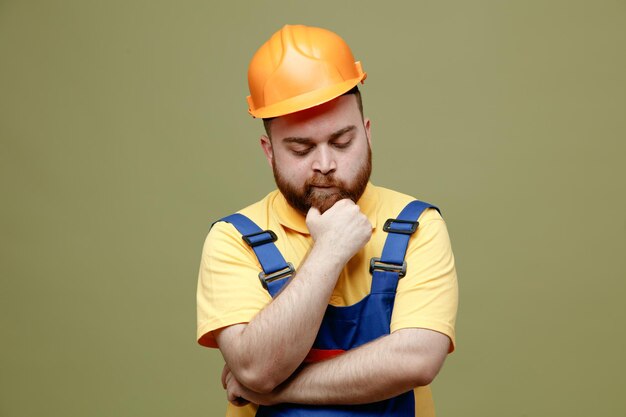 Pensando agarró la barbilla joven constructor hombre un uniforme usado en fondo verde