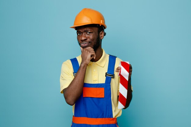 Pensando agarró la barbilla sosteniendo cinta adhesiva joven constructor afroamericano en uniforme aislado sobre fondo azul.