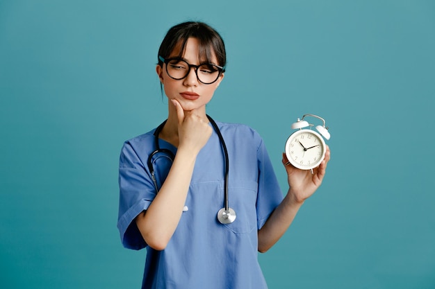 pensando agarró la barbilla sosteniendo el despertador joven doctora vistiendo uniforme fith estetoscopio aislado sobre fondo azul.